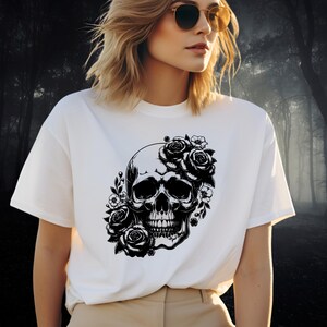 May include: White t-shirt featuring a black skull design adorned with roses and floral accents. The graphic is centered on the front of the shirt. The shirt has a crew neck and short sleeves.