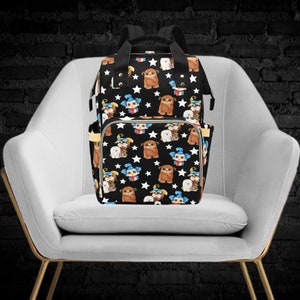 May include: A black diaper bag with a white star and cartoon character pattern. The bag has a large main compartment and a smaller front pocket. The bag is sitting on a white chair.