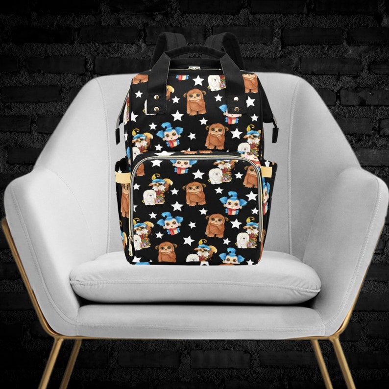 May include: A black diaper bag with a white star and cartoon character pattern. The bag has a large main compartment and a smaller front pocket. The bag is sitting on a white chair.