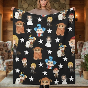 Può includere: Una coperta nera con un motivo ripetuto di personaggi dei cartoni animati e stelle bianche. I personaggi includono una creatura pelosa marrone, una donna in abito bianco e una creatura dalla pelle blu con una sciarpa rossa. La coperta è tenuta da una persona.
