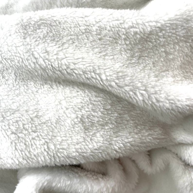 Pu&ograve; includere: Primo piano di un tessuto morbido, bianco e soffice. Il materiale ha un aspetto spesso e strutturato con un pelo denso e morbido. Le pieghe e le ombre creano un senso di profondit&agrave; e dimensione, suggerendo una coperta o un tessuto accogliente.