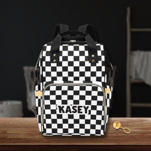 May include: Black and white checkered backpack with the name "Kasey" printed on the front pocket. The backpack has black straps and gold accents. A gold-colored rattle is on the wooden surface.
