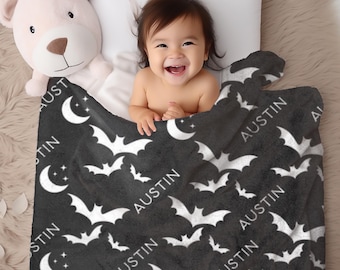 May include: A black baby blanket with a white bat and moon pattern. The name "AUSTIN" is printed in white. A smiling baby is under the blanket, next to a teddy bear.