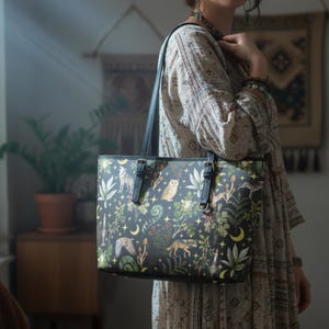 May include: Black tote bag featuring a forest-themed design with wolves, owls, and crescent moons. The bag has black handles and a black border. The forest scene includes green foliage and stars. The bag is carried over the shoulder.