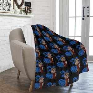 Pu&ograve; includere: Una coperta nera con un motivo ripetuto di un personaggio dei cartoni animati con capelli blu, una sciarpa rossa e un viso beige. La coperta &egrave; drappeggiata su una poltrona beige, con un interno bianco visibile.