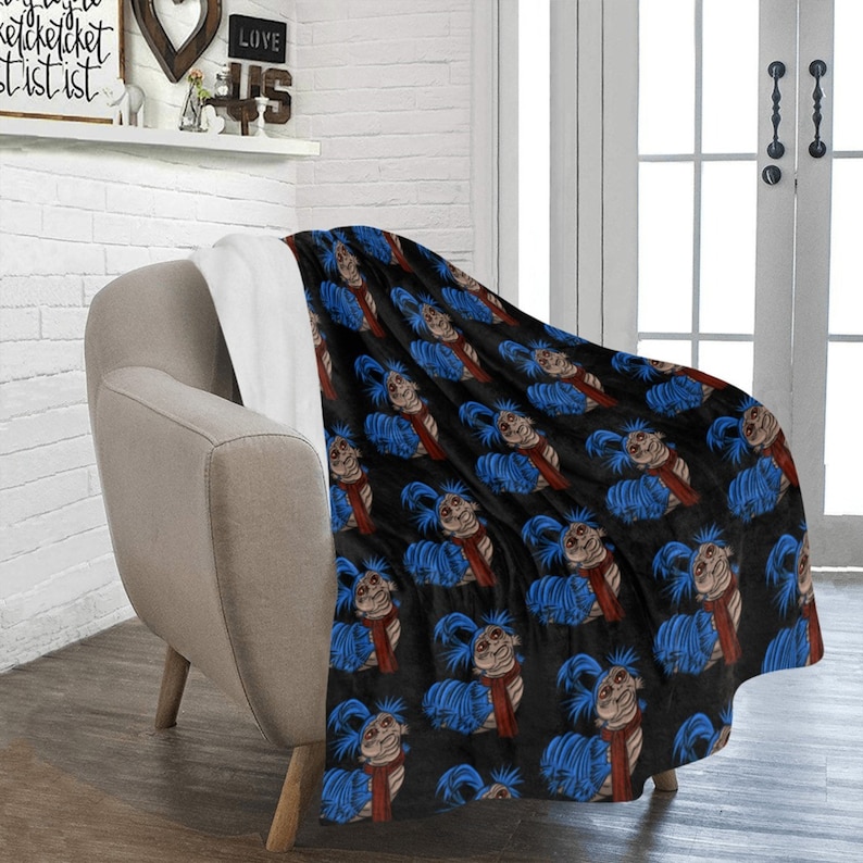 Pu&ograve; includere: Una coperta nera con un motivo ripetuto di un personaggio dei cartoni animati con capelli blu, una sciarpa rossa e un viso beige. La coperta &egrave; drappeggiata su una poltrona beige, con un interno bianco visibile.