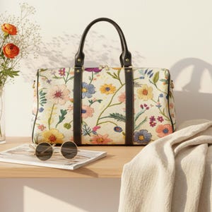 May include: A cream-coloured travel bag adorned with a floral pattern, complemented by black leather handles and details. The bag showcases a vibrant array of embroidered flowers. Sunglasses sit on a magazine on a wooden shelf, with a vase of flowers.