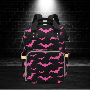 May include: Black backpack with a pink bat pattern. The bag has multiple compartments, a top handle, and adjustable shoulder straps. The exterior features a repeating pattern of pink bats against a black background, with gold-coloured zips and trim.