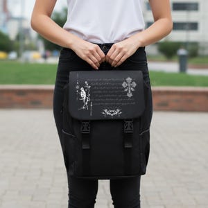 May include: Black backpack with a flap closure and two adjustable buckles. The flap features a white graphic design with a cross, script, and other decorative elements. The backpack is held by a person wearing a white shirt and black pants.
