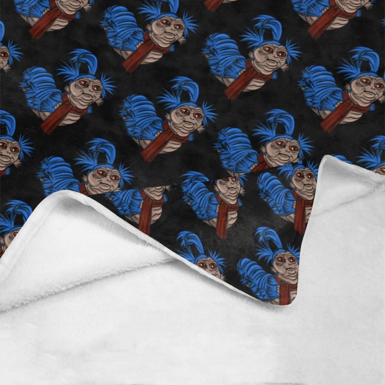 Pu&ograve; includere: Una coperta nera con un motivo ripetuto di un personaggio di verme dei cartoni animati con i capelli blu e una sciarpa rossa. La coperta ha un lato inferiore bianco e soffice.