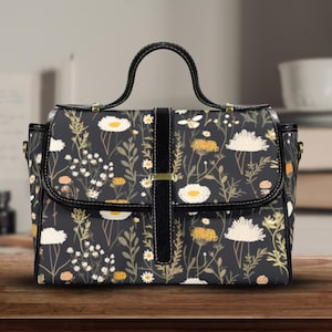 May include: A black satchel bag with a floral pattern featuring white daisies, yellow flowers, and green leaves. The bag has a black handle, a black strap, and gold-colored hardware. The bag is sitting on a wooden surface.