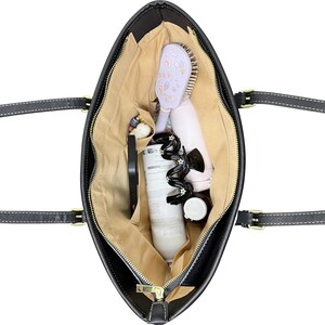 May include: Black tote bag with a gold zipper, filled with various items. Inside, a light purple hairbrush, a black phone, a white bottle, and a black hair accessory are visible. The bag has black handles with white stitching.
