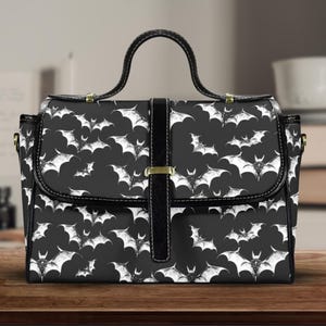 May include: Black handbag featuring a repeating pattern of white bats. The bag has a top handle, a flap closure, and a black strap with a gold-coloured buckle. The bag is on a wooden surface.