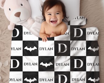 May include: A black and white checkered baby blanket with the name "DYLAN" and a bat symbol. The blanket is draped over a baby and a stuffed bear. The baby is smiling and looking at the camera.