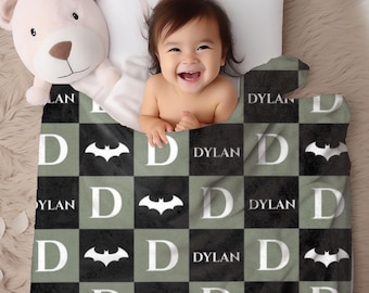 May include: A baby blanket with a black, gray, and white checkered pattern. The blanket features the name "DYLAN", the letter "D", and a bat symbol. A smiling baby is peeking over the blanket.