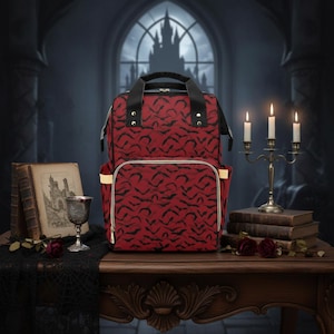 Puede incluir: Mochila roja con un patrón de murciélagos negros, asas negras y un bolsillo frontal con cremallera dorada. La bolsa está sobre una mesa de madera con un fondo de castillo gótico, velas y libros.