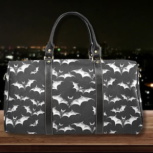 May include: A black duffel bag featuring a repeating white bat pattern. The bag includes two black leather-like handles and two vertical black straps running down the front, secured with gold-toned metal hardware at the base of the handles.