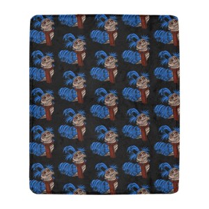 Pu&ograve; includere: Una coperta in peluche con un motivo ripetuto di una creatura blu a forma di verme con una sciarpa rossa. La creatura ha un viso marrone chiaro e capelli blu. Lo sfondo &egrave; nero.