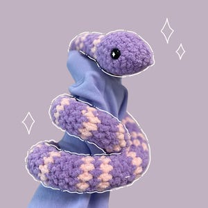 Op de afbeelding: Een handgemaakte gehaakte slang in tinten paars en crème. Het pluche speelgoed heeft een gedetailleerde kop met zwarte ogen en een patroon op het lichaam. De slang wordt getoond tegen een zachte paarse achtergrond.