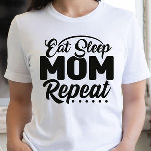 May include: White t-shirt with the text "Eat Sleep MOM Repeat" in black script and block letters. The shirt is paired with denim shorts. A blue and tan hat is visible in the background.