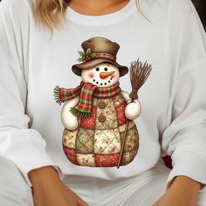 May include: White long-sleeved sweatshirt featuring a whimsical snowman graphic. The snowman wears a brown hat, a red and green plaid scarf, and holds a broom. The snowman's body is a patchwork design in red, green, and cream.