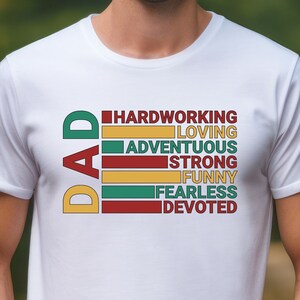 May include: White t-shirt with the word "DAD" in yellow, green, and red vertical text. Below, horizontal bars list qualities like "HARDWORKING," "LOVING," and "ADVENTUROUS" in a matching color scheme. The shirt is worn by a person.