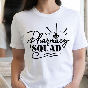 May include: A white short-sleeved t-shirt featuring the black text Pharmacy SQUAD printed on the front. The design includes a small pill graphic above the word Pharmacy and decorative lines underneath the word SQUAD, centered on the chest.