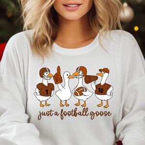 May include: White sweatshirt featuring a graphic of four cartoon geese in football gear. The geese wear helmets, jerseys with numbers, and hold a foam finger and a megaphone. The text "just a football goose" is below the geese.