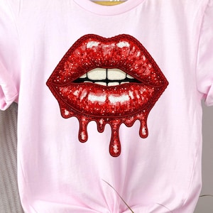 Op de afbeelding: Lichtroze t-shirt met een afbeelding van rode lippen met een druipend effect. De lippen hebben een pailletten textuur en witte tanden. Het shirt heeft korte mouwen en een knoop aan de onderkant.