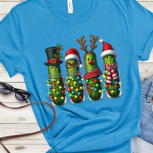 K&ouml;nnte beinhalten: Hellblaues T-Shirt mit einer skurrilen Illustration von vier Gew&uuml;rzgurken in Einmachgl&auml;sern, die jeweils mit festlichen Accessoires geschm&uuml;ckt sind. Die Gew&uuml;rzgurken sind mit Lichterketten, H&uuml;ten und Schals verziert.