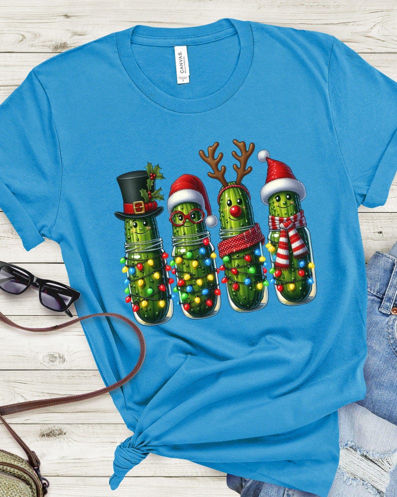 K&ouml;nnte beinhalten: Hellblaues T-Shirt mit einer skurrilen Illustration von vier Gew&uuml;rzgurken in Einmachgl&auml;sern, die jeweils mit festlichen Accessoires geschm&uuml;ckt sind. Die Gew&uuml;rzgurken sind mit Lichterketten, H&uuml;ten und Schals verziert.
