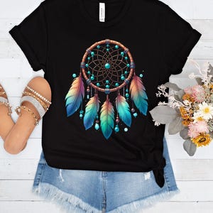 May include: Black t-shirt featuring a colorful dreamcatcher design. The dreamcatcher has a brown circular frame with a web of brown lines, turquoise beads, and colorful feathers in shades of blue, teal, and orange. The shirt is paired with denim shorts.