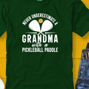 Op de afbeelding: Groen T-shirt met witte tekst die luidt: "Onderschat nooit een oma met een pickleball-bat". Twee witte pickleball-bats en een gele pickleball zijn afgebeeld boven de tekst.