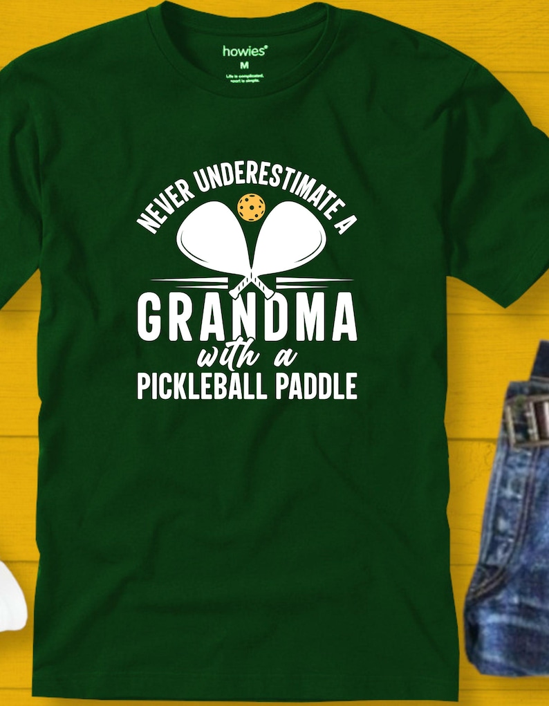 Op de afbeelding: Groen T-shirt met witte tekst die luidt: "Onderschat nooit een oma met een pickleball-bat". Twee witte pickleball-bats en een gele pickleball zijn afgebeeld boven de tekst.