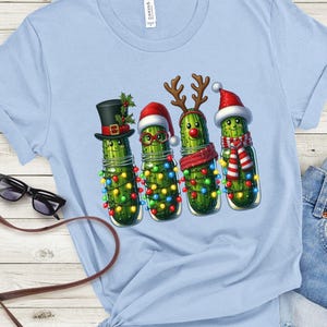 K&ouml;nnte beinhalten: Hellblaues T-Shirt mit einem weihnachtlichen Motiv. Das Design zeigt vier anthropomorphe Gurken in Gl&auml;sern, die jeweils mit festlichen Accessoires wie Zylinder, Weihnachtsm&uuml;tzen, Rentiergeweih und Schal geschm&uuml;ckt sind, alle mit Lichterketten.