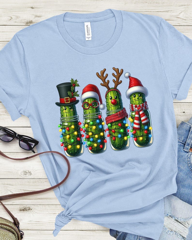K&ouml;nnte beinhalten: Hellblaues T-Shirt mit einem weihnachtlichen Motiv. Das Design zeigt vier anthropomorphe Gurken in Gl&auml;sern, die jeweils mit festlichen Accessoires wie Zylinder, Weihnachtsm&uuml;tzen, Rentiergeweih und Schal geschm&uuml;ckt sind, alle mit Lichterketten.