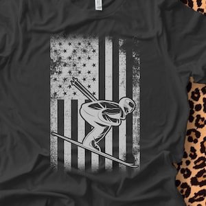 May include: Black t-shirt featuring a distressed American flag design with a skier in silhouette. The flag is in shades of gray and white, with a skier in action. The shirt is made of a soft material.