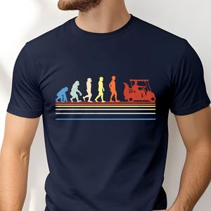 May include: Navy blue t-shirt featuring a colorful graphic of human evolution ending with a golf cart. The design includes figures in blue, teal, white, yellow, and orange, with horizontal stripes in the same colors. A fun tee for golf enthusiasts.