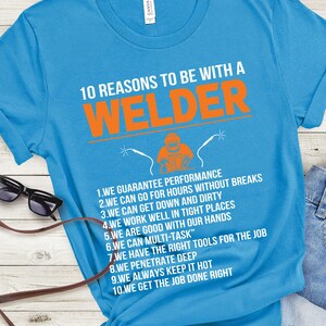May include: A turquoise t-shirt with the text "10 REASONS TO BE WITH A WELDER" in white and orange. The shirt features a graphic of a welder and a list of reasons. Sunglasses and a purse are visible.