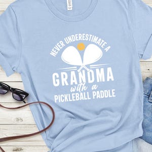 Op de afbeelding: Lichtblauw T-shirt met witte tekst die "Never underestimate a grandma with a pickleball paddle" luidt. Twee witte pickleball-rackets zijn boven de tekst afgebeeld met een gele pickleball.