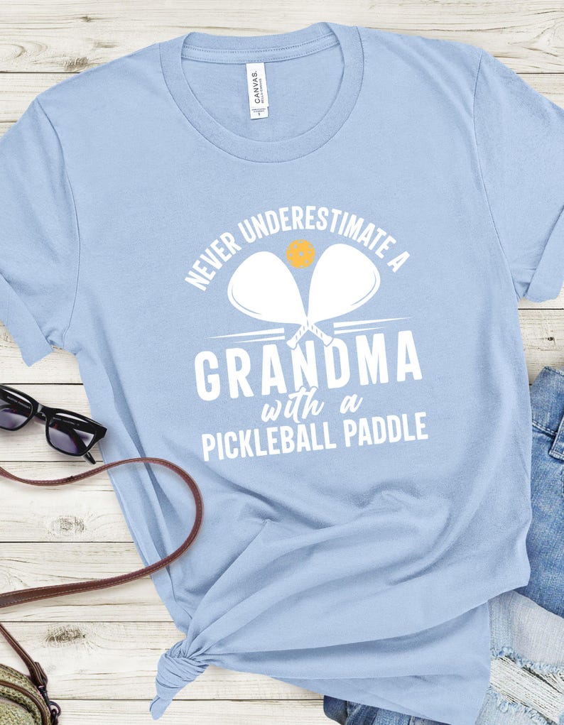 Op de afbeelding: Lichtblauw T-shirt met witte tekst die "Never underestimate a grandma with a pickleball paddle" luidt. Twee witte pickleball-rackets zijn boven de tekst afgebeeld met een gele pickleball.
