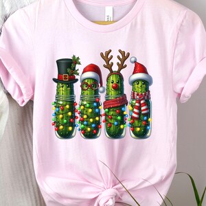 K&ouml;nnte beinhalten: Hellrosa T-Shirt mit einem festlichen Design. Das Motiv zeigt vier anthropomorphe Gurken in Gl&auml;sern, die jeweils mit Weihnachtsdekorationen geschm&uuml;ckt sind. Eine Gurke tr&auml;gt einen Zylinder, eine Brille, eine Rentiergeweih und die letzte einen Weihnachtsm&uuml;tze.