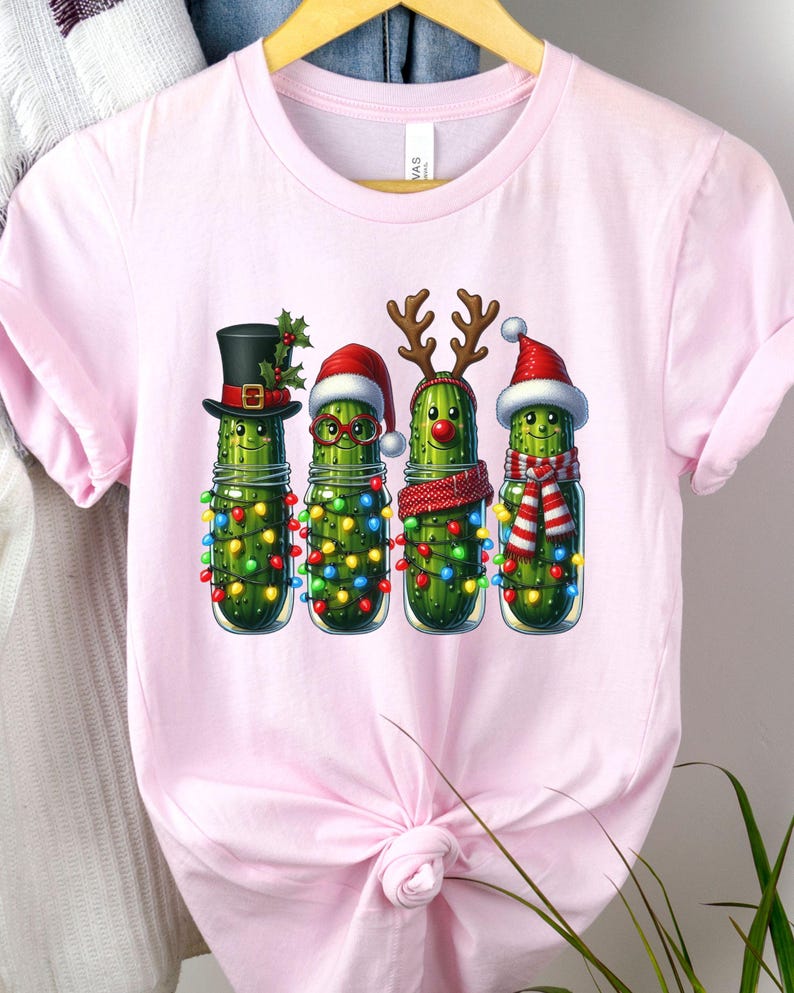 K&ouml;nnte beinhalten: Hellrosa T-Shirt mit einem festlichen Design. Das Motiv zeigt vier anthropomorphe Gurken in Gl&auml;sern, die jeweils mit Weihnachtsdekorationen geschm&uuml;ckt sind. Eine Gurke tr&auml;gt einen Zylinder, eine Brille, eine Rentiergeweih und die letzte einen Weihnachtsm&uuml;tze.