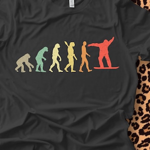May include: A black t-shirt with a vintage-style graphic of the evolution of man, culminating in a person snowboarding. The graphic is in shades of green, yellow, orange, and red.