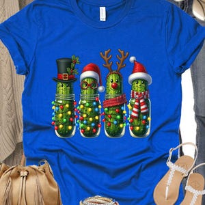 K&ouml;nnte beinhalten: K&ouml;nigsblaues T-Shirt mit einem weihnachtlichen Design. Das Design zeigt vier anthropomorphe Gurken in Gl&auml;sern, die jeweils mit festlichen Accessoires wie H&uuml;ten und Rentiergeweihen geschm&uuml;ckt sind. Die Gurken sind mit bunten Weihnachtslichtern verziert.