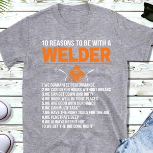 May include: A grey t-shirt with the text "10 REASONS TO BE WITH A WELDER" in orange and white. Below the text is a graphic of a welder. The shirt lists ten reasons to be with a welder.
