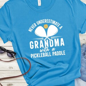 Op de afbeelding: Een lichtblauw T-shirt met de tekst "Never underestimate a grandma with a pickleball paddle" in wit. Twee witte pickleball-rackets zijn gekruist achter de tekst met een gele pickleball in het midden.