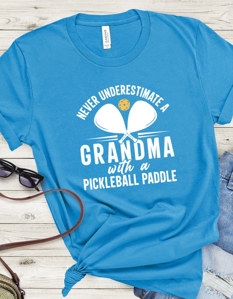 Op de afbeelding: Een lichtblauw T-shirt met de tekst "Never underestimate a grandma with a pickleball paddle" in wit. Twee witte pickleball-rackets zijn gekruist achter de tekst met een gele pickleball in het midden.