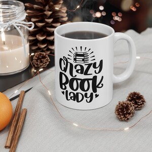 Peut inclure: Une tasse en céramique blanche avec un motif noir qui dit "Crazy Book Lady" avec une pile de livres et des cœurs. La tasse est remplie de café et repose sur une surface blanche avec une pomme de pin brune et des bâtonnets de cannelle.