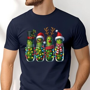 K&ouml;nnte beinhalten: Marineblaues T-Shirt mit einem weihnachtlichen Design. Die Grafik zeigt vier Gew&uuml;rzgurken in Gl&auml;sern, die jeweils mit festlichen Accessoires geschm&uuml;ckt sind: Zylinder, Weihnachtsm&uuml;tze, Rentiergeweih und Schal. Sie sind auch mit bunten Lichterketten verziert.